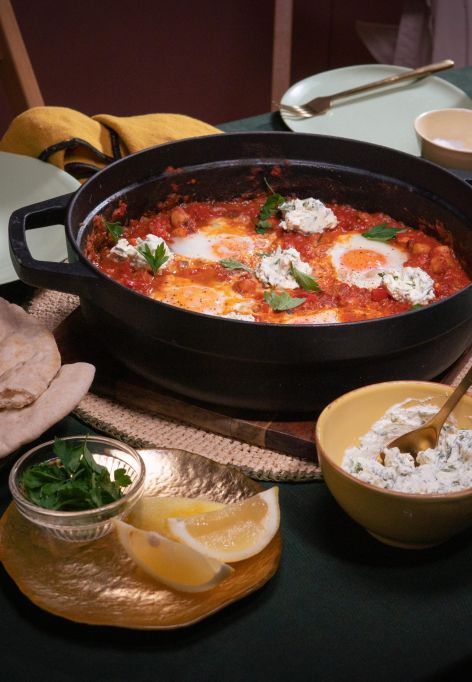 Tisch mit einer Pfanne Shakshuka – pochierte Eier in würziger Tomatensauce, garniert mit Kräutern und Klecksen von Frischkäse. Dazu Fladenbrot, Zitronenspalten und Kräuter.