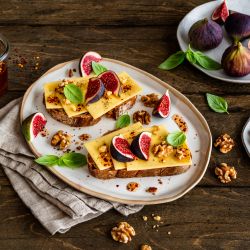 Zwei Scheiben Brot auf einem Teller, jeweils mit Butterkäse belegt, darauf frische Feigenstücke, geröstete Walnüsse und Basilikumblätter. Daneben ein Glas mit Chili-Honig und ein Holzlöffel, sowie zusätzliche Feigen und Walnüsse auf kleinen Tellern. Alles arrangiert auf einem rustikalen Holztisch.