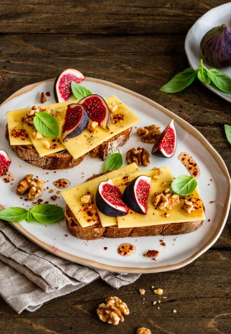 Zwei Scheiben Brot auf einem Teller, jeweils mit Butterkäse belegt, darauf frische Feigenstücke, geröstete Walnüsse und Basilikumblätter. Daneben ein Glas mit Chili-Honig und ein Holzlöffel, sowie zusätzliche Feigen und Walnüsse auf kleinen Tellern. Alles arrangiert auf einem rustikalen Holztisch.