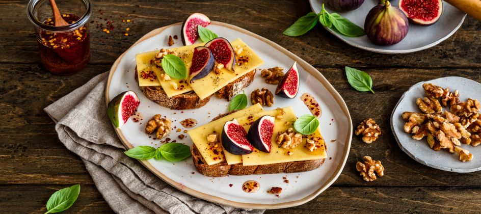 Zwei Scheiben Brot auf einem Teller, jeweils mit Butterkäse belegt, darauf frische Feigenstücke, geröstete Walnüsse und Basilikumblätter. Daneben ein Glas mit Chili-Honig und ein Holzlöffel, sowie zusätzliche Feigen und Walnüsse auf kleinen Tellern. Alles arrangiert auf einem rustikalen Holztisch.