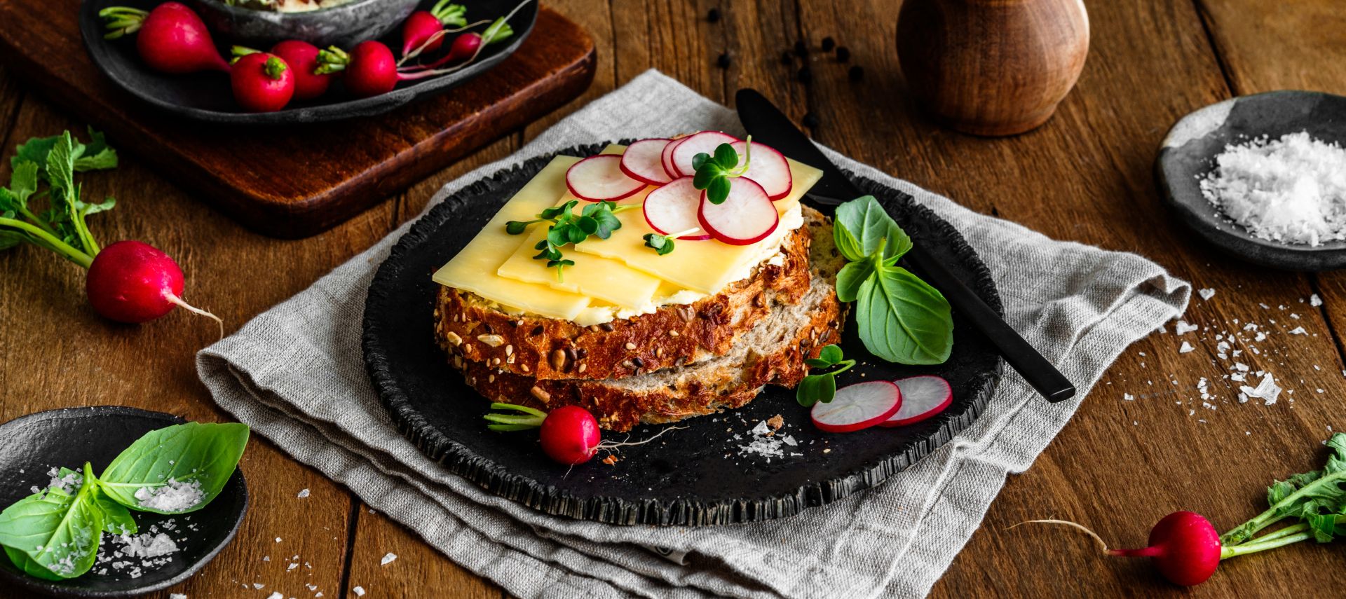 Eine Scheibe Körnerbrot auf einem schwarzen Teller, belegt mit würzigem Cheddar und dünn geschnittenen Radieschenscheiben, garniert mit frischen Kräutern und Basilikumblättern. Rundherum liegen einzelne Radieschen, Salzflocken und ein kleiner Bund Kräuter. Im Hintergrund ein Holzbrett mit Kresse und Radieschen sowie eine Pfeffermühle.