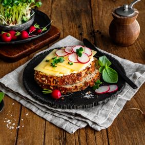 Eine Scheibe Körnerbrot auf einem schwarzen Teller, belegt mit würzigem Cheddar und dünn geschnittenen Radieschenscheiben, garniert mit frischen Kräutern und Basilikumblättern. Rundherum liegen einzelne Radieschen, Salzflocken und ein kleiner Bund Kräuter. Im Hintergrund ein Holzbrett mit Kresse und Radieschen sowie eine Pfeffermühle.