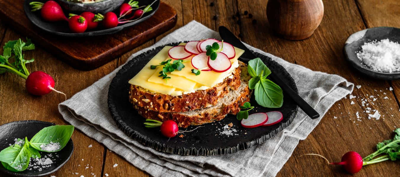 Eine Scheibe Körnerbrot auf einem schwarzen Teller, belegt mit würzigem Cheddar und dünn geschnittenen Radieschenscheiben, garniert mit frischen Kräutern und Basilikumblättern. Rundherum liegen einzelne Radieschen, Salzflocken und ein kleiner Bund Kräuter. Im Hintergrund ein Holzbrett mit Kresse und Radieschen sowie eine Pfeffermühle.