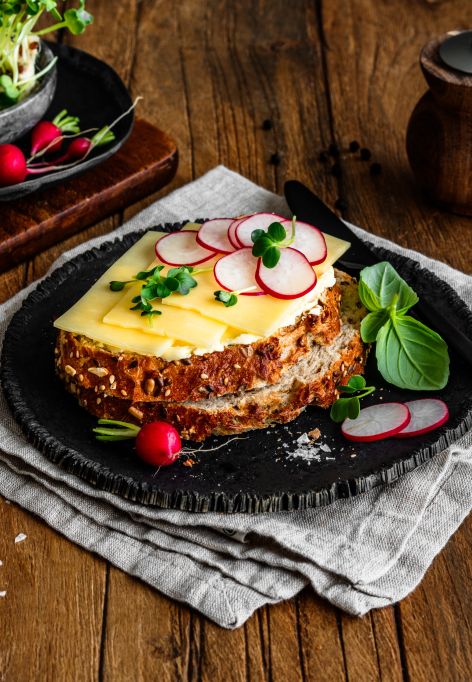 Eine Scheibe Körnerbrot auf einem schwarzen Teller, belegt mit würzigem Cheddar und dünn geschnittenen Radieschenscheiben, garniert mit frischen Kräutern und Basilikumblättern. Rundherum liegen einzelne Radieschen, Salzflocken und ein kleiner Bund Kräuter. Im Hintergrund ein Holzbrett mit Kresse und Radieschen sowie eine Pfeffermühle.