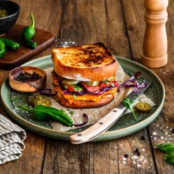 Geröstetes Sandwich aus zwei Scheiben Kastenweißbrot, gefüllt mit geschmolzenem Chili-Cheddar, gebratener Chorizo, roten Zwiebelringen, Jalapeños und frischen Kräutern. Serviert auf einem grünen Teller mit einem Messer, umgeben von Salzflocken, Pfefferkörnern, grünen Chilis und einer Pfeffermühle auf einem rustikalen Holztisch.