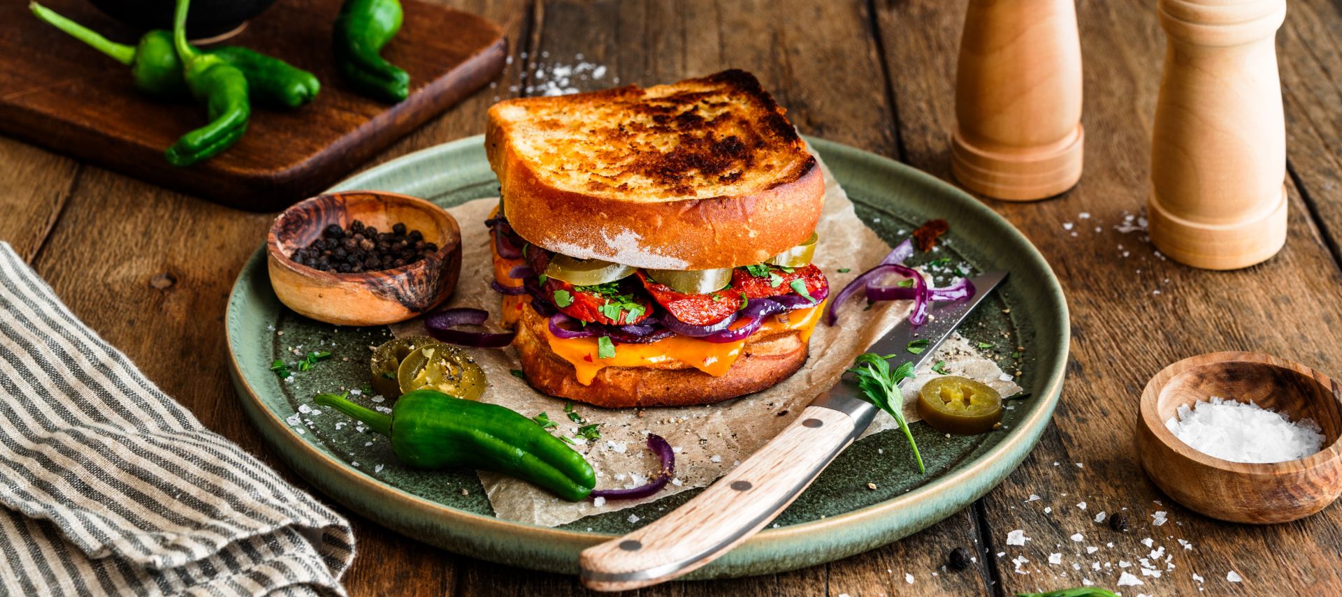 Geröstetes Sandwich aus zwei Scheiben Kastenweißbrot, gefüllt mit geschmolzenem Chili-Cheddar, gebratener Chorizo, roten Zwiebelringen, Jalapeños und frischen Kräutern. Serviert auf einem grünen Teller mit einem Messer, umgeben von Salzflocken, Pfefferkörnern, grünen Chilis und einer Pfeffermühle auf einem rustikalen Holztisch.