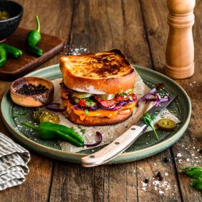 Geröstetes Sandwich aus zwei Scheiben Kastenweißbrot, gefüllt mit geschmolzenem Chili-Cheddar, gebratener Chorizo, roten Zwiebelringen, Jalapeños und frischen Kräutern. Serviert auf einem grünen Teller mit einem Messer, umgeben von Salzflocken, Pfefferkörnern, grünen Chilis und einer Pfeffermühle auf einem rustikalen Holztisch.