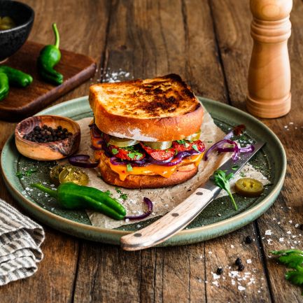 Geröstetes Sandwich aus zwei Scheiben Kastenweißbrot, gefüllt mit geschmolzenem Chili-Cheddar, gebratener Chorizo, roten Zwiebelringen, Jalapeños und frischen Kräutern. Serviert auf einem grünen Teller mit einem Messer, umgeben von Salzflocken, Pfefferkörnern, grünen Chilis und einer Pfeffermühle auf einem rustikalen Holztisch.