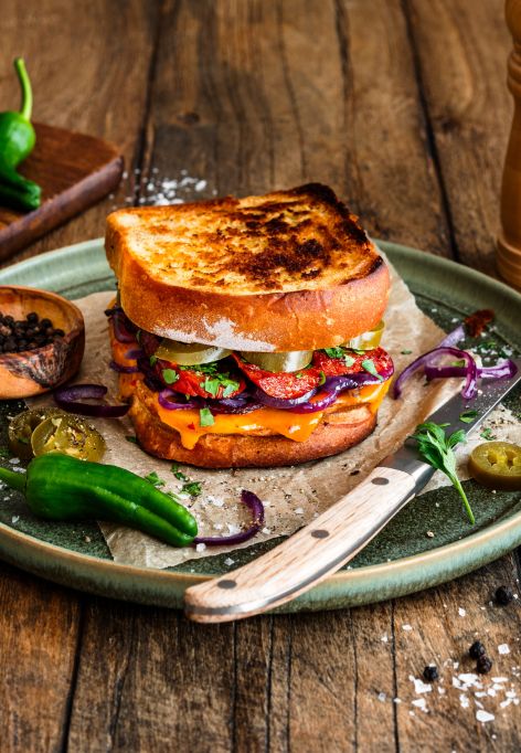 Geröstetes Sandwich aus zwei Scheiben Kastenweißbrot, gefüllt mit geschmolzenem Chili-Cheddar, gebratener Chorizo, roten Zwiebelringen, Jalapeños und frischen Kräutern. Serviert auf einem grünen Teller mit einem Messer, umgeben von Salzflocken, Pfefferkörnern, grünen Chilis und einer Pfeffermühle auf einem rustikalen Holztisch.