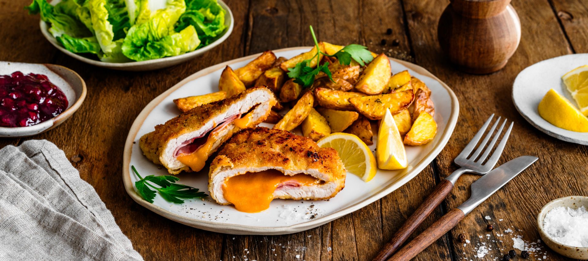 Ein Teller mit knusprigem Cordon Bleu, aufgeschnitten und gefüllt mit geschmolzenem Käse und Schinken, serviert mit goldbraunen Kartoffelspalten und Zitronenspalten. Daneben liegen eine Gabel und ein Messer auf einem Holztisch.
