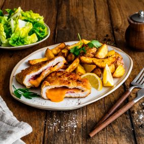 Ein Teller mit knusprigem Cordon Bleu, aufgeschnitten und gefüllt mit geschmolzenem Käse und Schinken, serviert mit goldbraunen Kartoffelspalten und Zitronenspalten. Daneben liegen eine Gabel und ein Messer auf einem Holztisch.