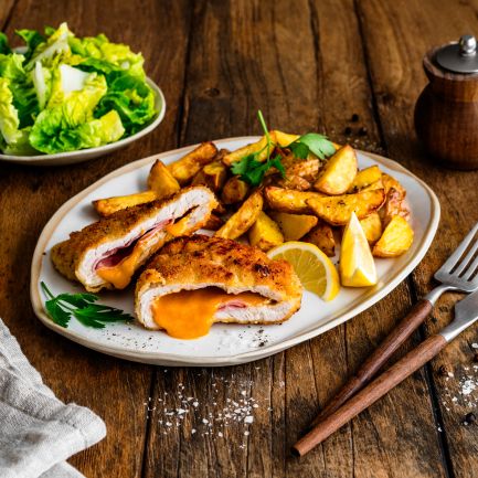 Ein Teller mit knusprigem Cordon Bleu, aufgeschnitten und gefüllt mit geschmolzenem Käse und Schinken, serviert mit goldbraunen Kartoffelspalten und Zitronenspalten. Daneben liegen eine Gabel und ein Messer auf einem Holztisch.