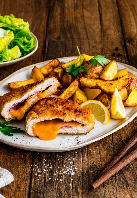 Ein Teller mit knusprigem Cordon Bleu, aufgeschnitten und gefüllt mit geschmolzenem Käse und Schinken, serviert mit goldbraunen Kartoffelspalten und Zitronenspalten. Daneben liegen eine Gabel und ein Messer auf einem Holztisch.