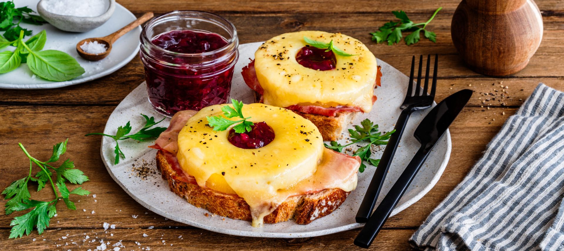 Zwei Toast-Hawaii-Scheiben auf einem Teller, jeweils belegt mit Schinken, geschmolzenem Käse, einer Ananasscheibe und einem Klecks Preiselbeeren, garniert mit Petersilie. Daneben ein Glas Preiselbeeren, frische Kräuter, Salzschale und eine Pfeffermühle auf einem rustikalen Holztisch.