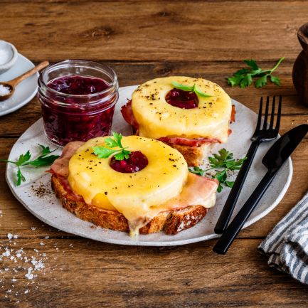 Zwei Toast-Hawaii-Scheiben auf einem Teller, jeweils belegt mit Schinken, geschmolzenem Käse, einer Ananasscheibe und einem Klecks Preiselbeeren, garniert mit Petersilie. Daneben ein Glas Preiselbeeren, frische Kräuter, Salzschale und eine Pfeffermühle auf einem rustikalen Holztisch.