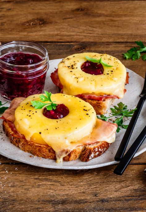Zwei Toast-Hawaii-Scheiben auf einem Teller, jeweils belegt mit Schinken, geschmolzenem Käse, einer Ananasscheibe und einem Klecks Preiselbeeren, garniert mit Petersilie. Daneben ein Glas Preiselbeeren, frische Kräuter, Salzschale und eine Pfeffermühle auf einem rustikalen Holztisch.