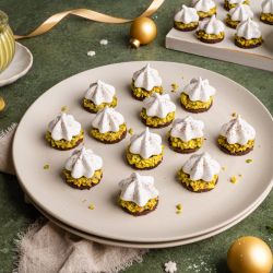 Ein Teller mit festlich dekorierten Pistazien-Plätzchen: kleine runde Kekse mit grünen gehackten Pistazien, darauf weiße Baiser-Tupfen, leicht mit Kakaopulver bestäubt. Im Hintergrund weitere Plätzchen, ein Glas Pistaziencreme und goldene Weihnachtskugeln auf grünem Untergrund.