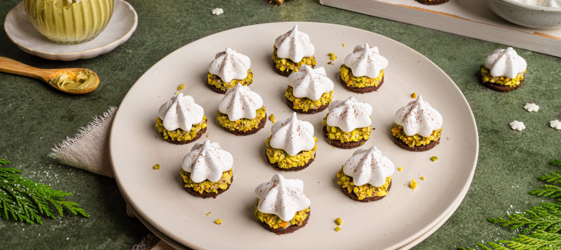 Ein Teller mit festlich dekorierten Pistazien-Plätzchen: kleine runde Kekse mit grünen gehackten Pistazien, darauf weiße Baiser-Tupfen, leicht mit Kakaopulver bestäubt. Im Hintergrund weitere Plätzchen, ein Glas Pistaziencreme und goldene Weihnachtskugeln auf grünem Untergrund.