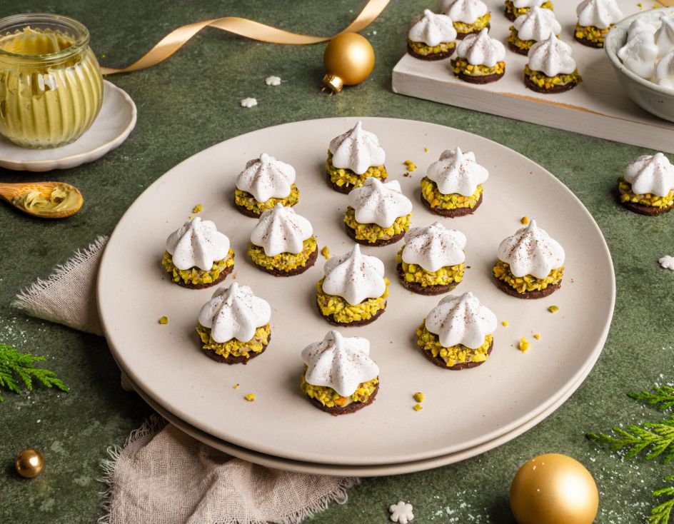 Ein Teller mit festlich dekorierten Pistazien-Plätzchen: kleine runde Kekse mit grünen gehackten Pistazien, darauf weiße Baiser-Tupfen, leicht mit Kakaopulver bestäubt. Im Hintergrund weitere Plätzchen, ein Glas Pistaziencreme und goldene Weihnachtskugeln auf grünem Untergrund.