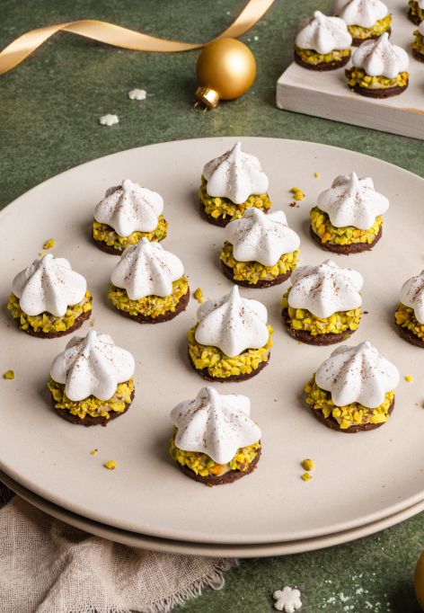 Ein Teller mit festlich dekorierten Pistazien-Plätzchen: kleine runde Kekse mit grünen gehackten Pistazien, darauf weiße Baiser-Tupfen, leicht mit Kakaopulver bestäubt. Im Hintergrund weitere Plätzchen, ein Glas Pistaziencreme und goldene Weihnachtskugeln auf grünem Untergrund.