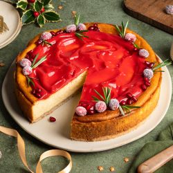 Festlich dekorierter Käsekuchen auf einem Teller: goldbrauner Kuchen mit einer glänzenden roten Cranberry-Punsch-Curd-Schicht, garniert mit gezuckerten Cranberries, Rosmarinzweigen und Granatapfelkernen. Daneben eine Schale mit Cranberries, goldene Schleifen und weihnachtliche Dekoration auf grünem Untergrund.