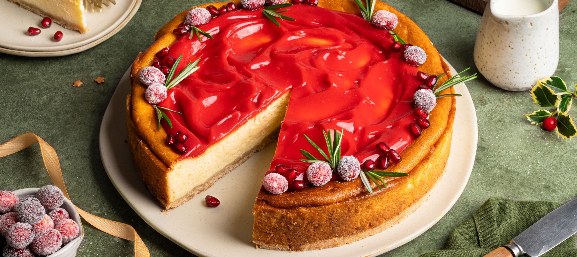 Festlich dekorierter Käsekuchen auf einem Teller: goldbrauner Kuchen mit einer glänzenden roten Cranberry-Punsch-Curd-Schicht, garniert mit gezuckerten Cranberries, Rosmarinzweigen und Granatapfelkernen. Daneben eine Schale mit Cranberries, goldene Schleifen und weihnachtliche Dekoration auf grünem Untergrund.