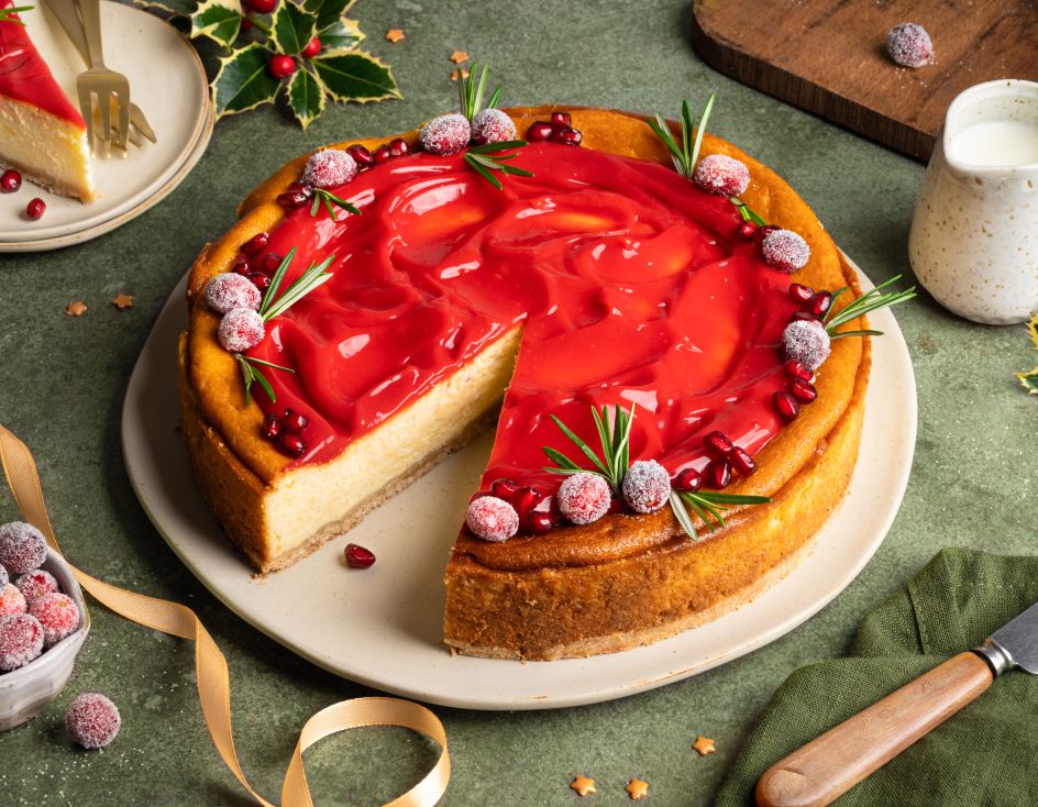 Festlich dekorierter Käsekuchen auf einem Teller: goldbrauner Kuchen mit einer glänzenden roten Cranberry-Punsch-Curd-Schicht, garniert mit gezuckerten Cranberries, Rosmarinzweigen und Granatapfelkernen. Daneben eine Schale mit Cranberries, goldene Schleifen und weihnachtliche Dekoration auf grünem Untergrund.