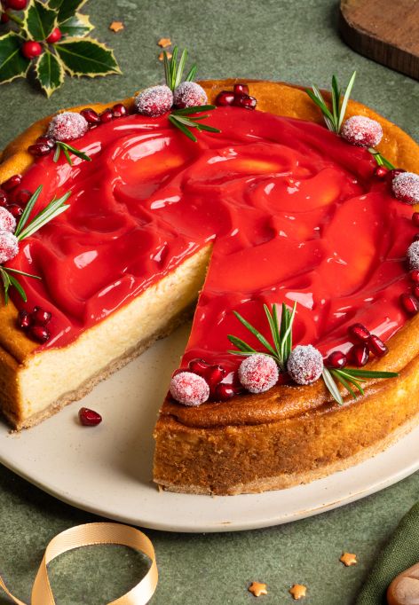 Festlich dekorierter Käsekuchen auf einem Teller: goldbrauner Kuchen mit einer glänzenden roten Cranberry-Punsch-Curd-Schicht, garniert mit gezuckerten Cranberries, Rosmarinzweigen und Granatapfelkernen. Daneben eine Schale mit Cranberries, goldene Schleifen und weihnachtliche Dekoration auf grünem Untergrund.