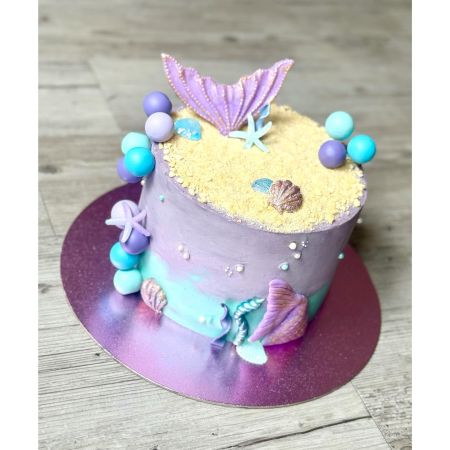 Runde Motivtorte mit Meerjungfrauen-Design: Die Torte ist in Lila- und Türkistönen gestaltet, mit essbaren Perlen, Muscheln und einer großen lilafarbenen Meerjungfrauenflosse als Dekoration. Der obere Teil ist mit zerbröselten Keksen als Sand bedeckt, umgeben von bunten Kugeln in Blau- und Lilatönen. Die Torte steht auf einer glänzenden, pink-violetten Tortenplatte.