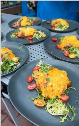 Mehrere schwarze Teller mit einer herzhaften Portion Shepherd’s Pie-Variation: überbackenes Kartoffel-Kürbis-Püree mit goldbrauner Käsekruste, darunter Wildragout und Champignons. Daneben frischer Salat mit grünen Blättern und halbierten Kirschtomaten. Die Teller stehen auf einem Metalltisch, angerichtet für mehrere Personen.