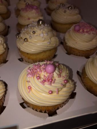 Mehrere Cupcakes in einer weißen Transportbox, dekoriert mit cremigem, hellgelbem Frosting und rosa sowie silbernen Zuckerperlen. Die Cupcakes sind gleichmäßig angeordnet, einige mit rosa Streuseln und großen Perlen, andere mit weißen und silbernen Dekorationen.