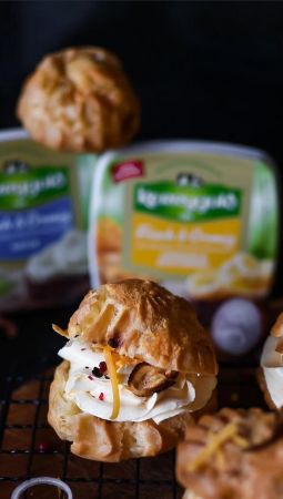 Nahaufnahme eines gefüllten Brandteigs (Profiterole) auf einem schwarzen Gitter: Die Füllung besteht aus cremigem Belag, garniert mit Zwiebelscheiben, geriebenen Cheddar und roten Pfefferstückchen.