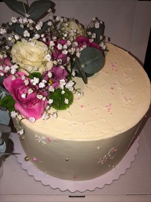 Runde Torte mit glatter, cremefarbener Oberfläche, dekoriert mit frischen Blumen: weiße und pinke Rosen, kleine weiße Blüten und grüne Blätter. Die Torte steht auf einer weißen Tortenscheibe und ist mit feinen rosa und weißen Zuckerstreuseln verziert.