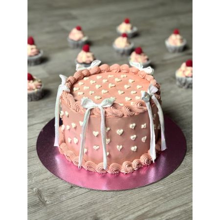 Runde rosa Torte auf einer glänzenden pinken Tortenplatte, dekoriert mit weißen kleinen Herzmustern, geschwungenem Rand aus rosa Creme und vier weißen Schleifen an den Seiten. Im Hintergrund mehrere kleine Cupcakes mit rosa und weißem Frosting und einer Himbeere als Topping.