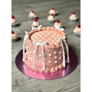 Runde rosa Torte auf einer glänzenden pinken Tortenplatte, dekoriert mit weißen kleinen Herzmustern, geschwungenem Rand aus rosa Creme und vier weißen Schleifen an den Seiten. Im Hintergrund mehrere kleine Cupcakes mit rosa und weißem Frosting und einer Himbeere als Topping.