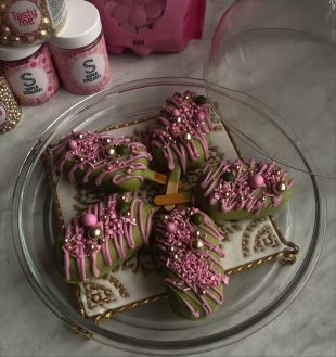 Fünf Pistazien-Matcha-Cakesicles auf einem Glas-Teller: grüne Kuchen am Stiel, dekoriert mit rosa Schokoladenguss, rosa und goldenen Zuckerperlen sowie Streuseln. Im Hintergrund stehen rosa und goldene Dekorationsstreusel in Dosen.