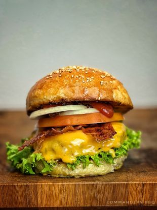 Nahaufnahme eines saftigen Burgers auf einem Holzbrett: Sesambrötchen, darunter frischer grüner Salat, geschmolzener Cheddar-Käse, knuspriger Bacon, Tomatenscheibe, Zwiebelringe und etwas Ketchup. Der Burger wirkt frisch und appetitlich angerichtet.