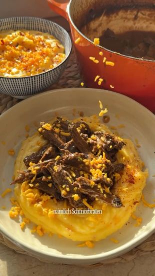 Teller mit cremigem Kartoffel-Cheddar-Püree, darauf zartes Schmorbratenfleisch in Streifen, garniert mit geriebenem Käse und etwas Paprikapulver. Daneben eine Schüssel mit zusätzlichem Püree und ein orangefarbener Schmortopf im Hintergrund