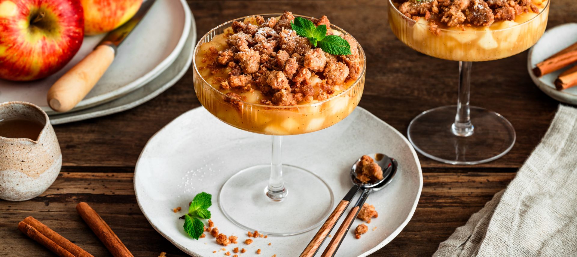 Zwei Gläser mit Apple Crumble im Glas: unten geschichtete Apfelwürfel, oben goldbraune Streusel, garniert mit Minzblättern. Daneben liegen Zimtstangen, ein kleiner Keramikkrug und rote Äpfel auf einem Teller auf einem rustikalen Holztisch.
