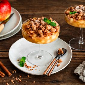 Zwei Gläser mit Apple Crumble im Glas: unten geschichtete Apfelwürfel, oben goldbraune Streusel, garniert mit Minzblättern. Daneben liegen Zimtstangen, ein kleiner Keramikkrug und rote Äpfel auf einem Teller auf einem rustikalen Holztisch.