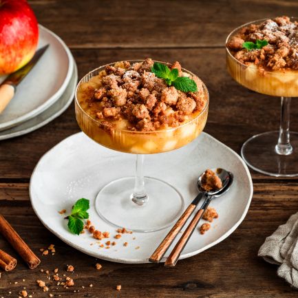 Zwei Gläser mit Apple Crumble im Glas: unten geschichtete Apfelwürfel, oben goldbraune Streusel, garniert mit Minzblättern. Daneben liegen Zimtstangen, ein kleiner Keramikkrug und rote Äpfel auf einem Teller auf einem rustikalen Holztisch.