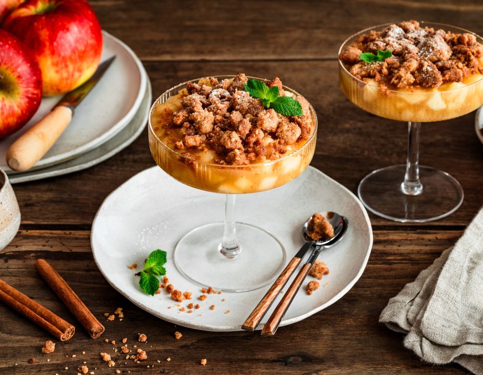 Zwei Gläser mit Apple Crumble im Glas: unten geschichtete Apfelwürfel, oben goldbraune Streusel, garniert mit Minzblättern. Daneben liegen Zimtstangen, ein kleiner Keramikkrug und rote Äpfel auf einem Teller auf einem rustikalen Holztisch.