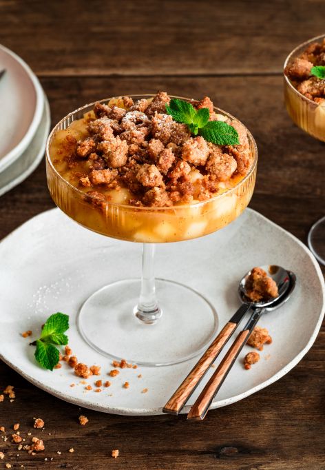 Zwei Gläser mit Apple Crumble im Glas: unten geschichtete Apfelwürfel, oben goldbraune Streusel, garniert mit Minzblättern. Daneben liegen Zimtstangen, ein kleiner Keramikkrug und rote Äpfel auf einem Teller auf einem rustikalen Holztisch.