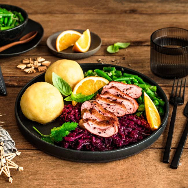 Festlich angerichteter Teller mit rosa gebratener Entenbrust in Scheiben, Rotkohl, zwei Kartoffelklößen und grünen Bohnen, garniert mit Orangenspalten und frischen Kräutern. Im Hintergrund liegen weitere Bohnen in einer Schale, Orangenspalten auf einem kleinen Teller sowie weihnachtliche Dekoration auf einem rustikalen Holztisch.