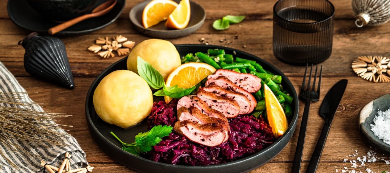 Festlich angerichteter Teller mit rosa gebratener Entenbrust in Scheiben, Rotkohl, zwei Kartoffelklößen und grünen Bohnen, garniert mit Orangenspalten und frischen Kräutern. Im Hintergrund liegen weitere Bohnen in einer Schale, Orangenspalten auf einem kleinen Teller sowie weihnachtliche Dekoration auf einem rustikalen Holztisch.