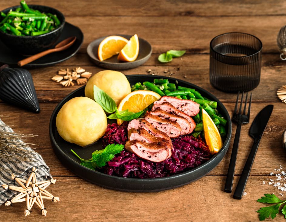 Festlich angerichteter Teller mit rosa gebratener Entenbrust in Scheiben, Rotkohl, zwei Kartoffelklößen und grünen Bohnen, garniert mit Orangenspalten und frischen Kräutern. Im Hintergrund liegen weitere Bohnen in einer Schale, Orangenspalten auf einem kleinen Teller sowie weihnachtliche Dekoration auf einem rustikalen Holztisch.