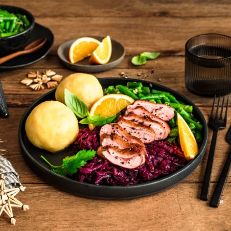 Festlich angerichteter Teller mit rosa gebratener Entenbrust in Scheiben, Rotkohl, zwei Kartoffelklößen und grünen Bohnen, garniert mit Orangenspalten und frischen Kräutern. Im Hintergrund liegen weitere Bohnen in einer Schale, Orangenspalten auf einem kleinen Teller sowie weihnachtliche Dekoration auf einem rustikalen Holztisch.