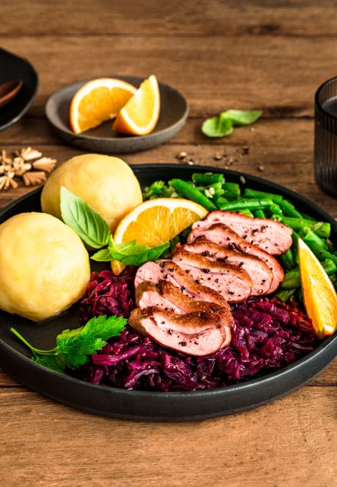 Festlich angerichteter Teller mit rosa gebratener Entenbrust in Scheiben, Rotkohl, zwei Kartoffelklößen und grünen Bohnen, garniert mit Orangenspalten und frischen Kräutern. Im Hintergrund liegen weitere Bohnen in einer Schale, Orangenspalten auf einem kleinen Teller sowie weihnachtliche Dekoration auf einem rustikalen Holztisch.