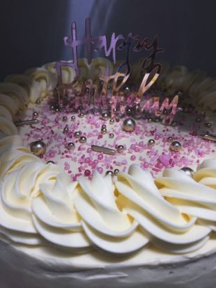 Runder Geburtstagskuchen mit weißer Sahnecreme am Rand, dekoriert mit rosa Zuckerstreuseln und silbernen Perlen. In der Mitte steckt ein glänzender Schriftzug ‚Happy Birthday‘.