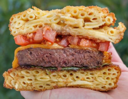 Nahaufnahme eines Burgers. Das Brötchen besteht aus frittierten Makkaroni-Nudeln. Zwischen den Nudelschichten befinden sich ein saftiges Rindfleisch-Patty, geschmolzener Cheddar-Käse, frische Tomatenwürfel und etwas Salat.